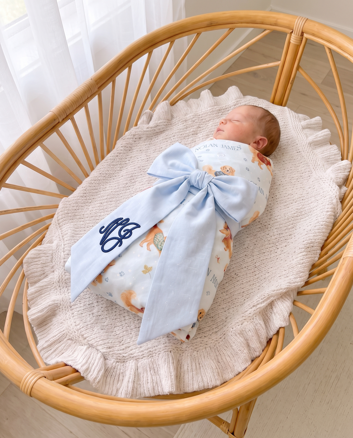 Swaddled newborn in a rattan bassinet with the light blue bow sash and navy script monogram