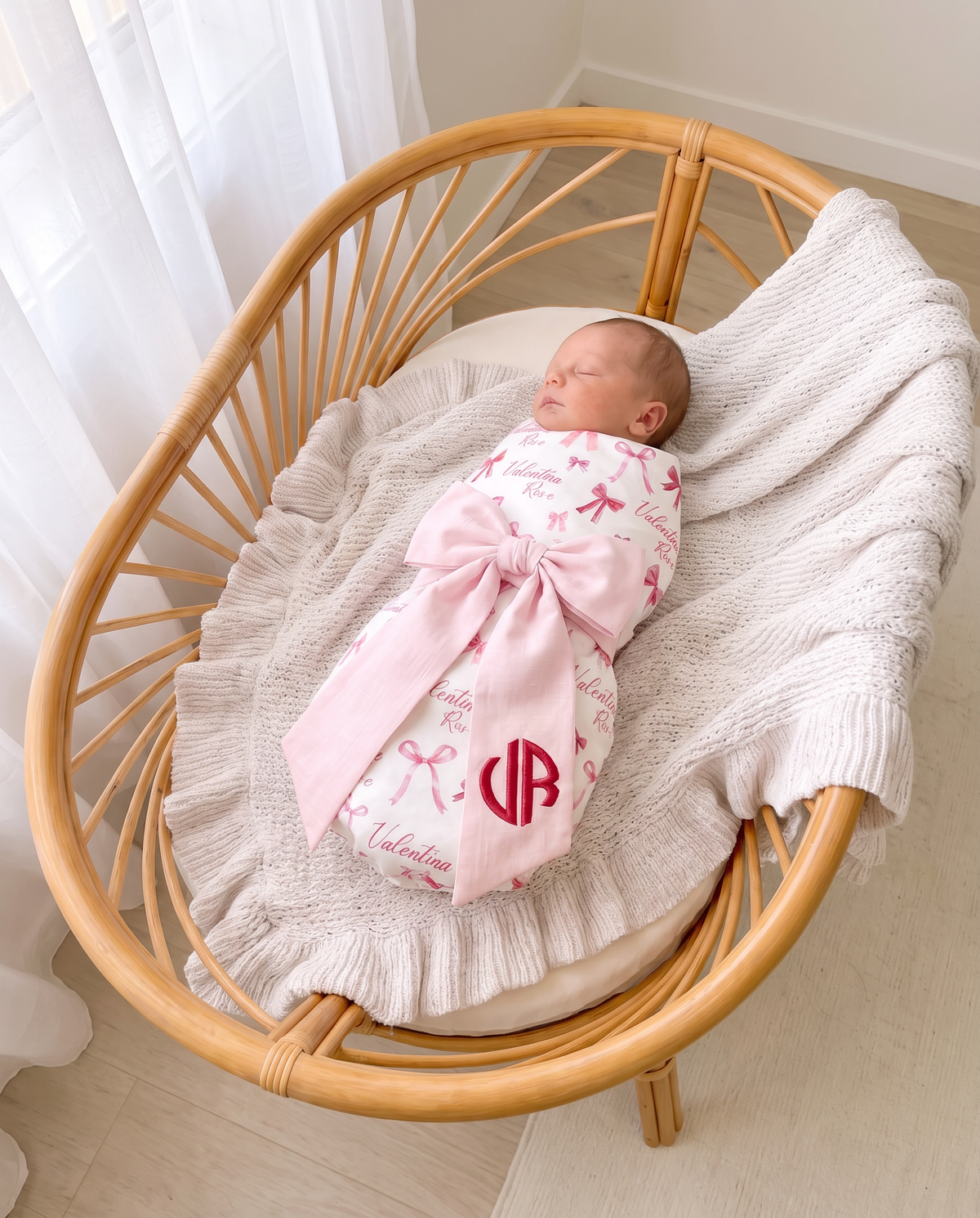 Swaddled newborn in a rattan bassinet with the light pink bow sash tied across