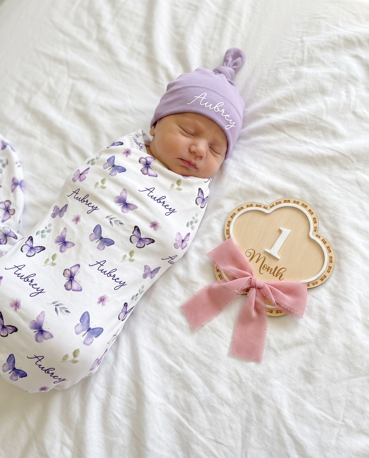 Flowers monthly milestone wood sign set to 1 Month beside a swaddled newborn on a white bed