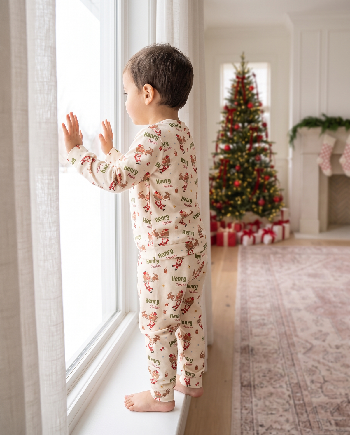 Toddler in Santa's Joyride pajamas looking out the window by the Christmas tree