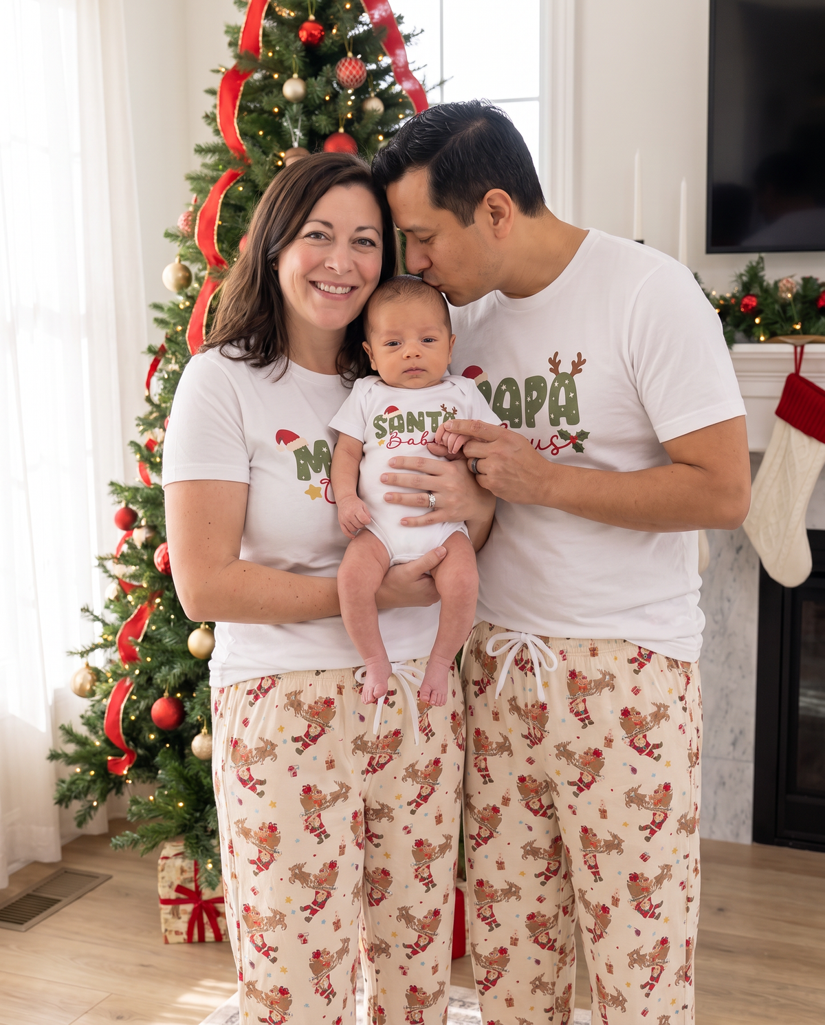 Parents in Claus graphic tees holding their baby in front of the Christmas tree