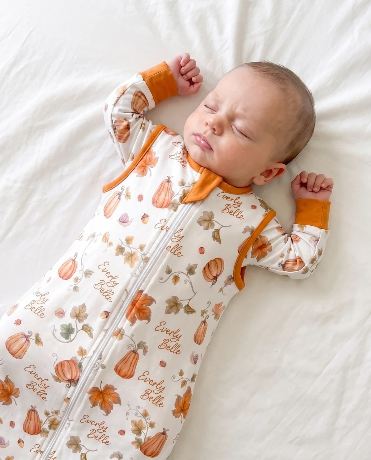 A baby in a personalized sleep bag printed with pumpkins, acorns and autumn leaves, the name Everly Belle repeated across the fabric