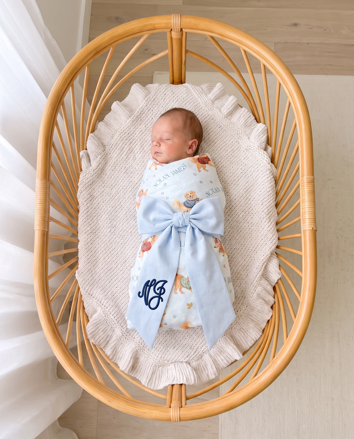 Newborn swaddled in Winter Wag print sleeping in a rattan bassinet with a light blue monogrammed bow sash