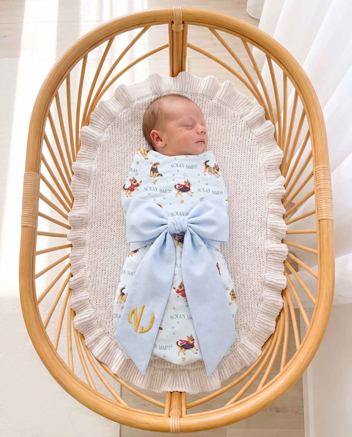 Newborn in a rattan bassinet with the light blue bow sash and gold round monogram