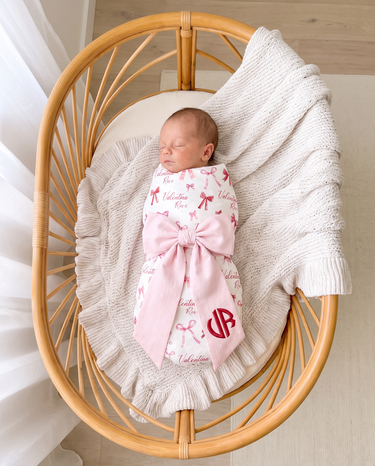 Newborn swaddled in White Coquette print sleeping in a rattan bassinet with a light pink monogrammed bow sash