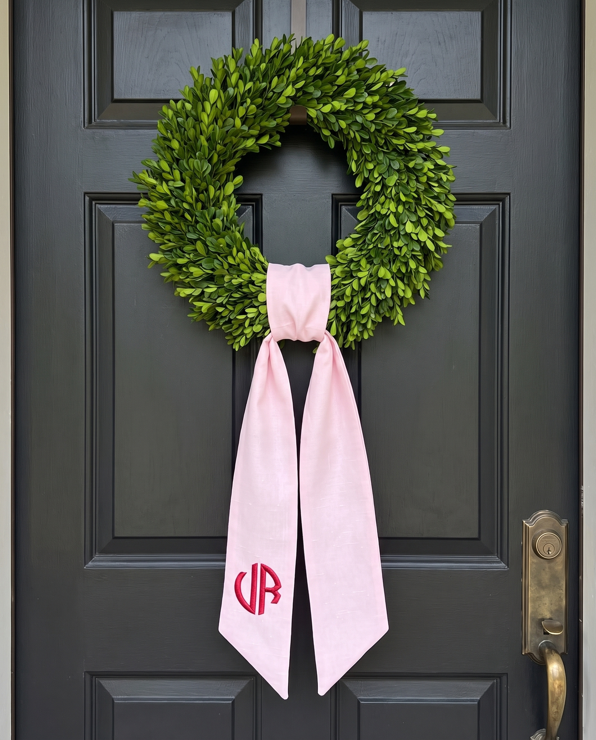 Light pink monogrammed bow sash hanging on a dark front door with a green wreath