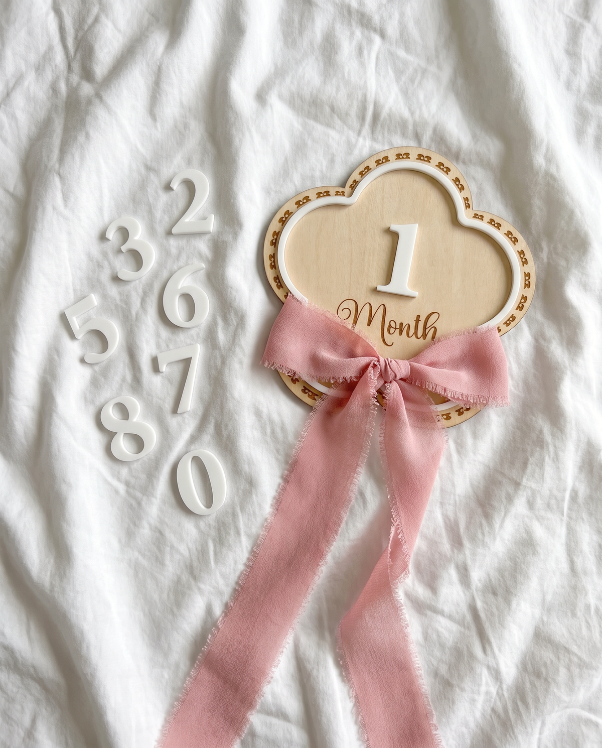 Flowers monthly milestone wood sign with a pink ribbon bow and a set of white acrylic numbers laid out on a white blanket