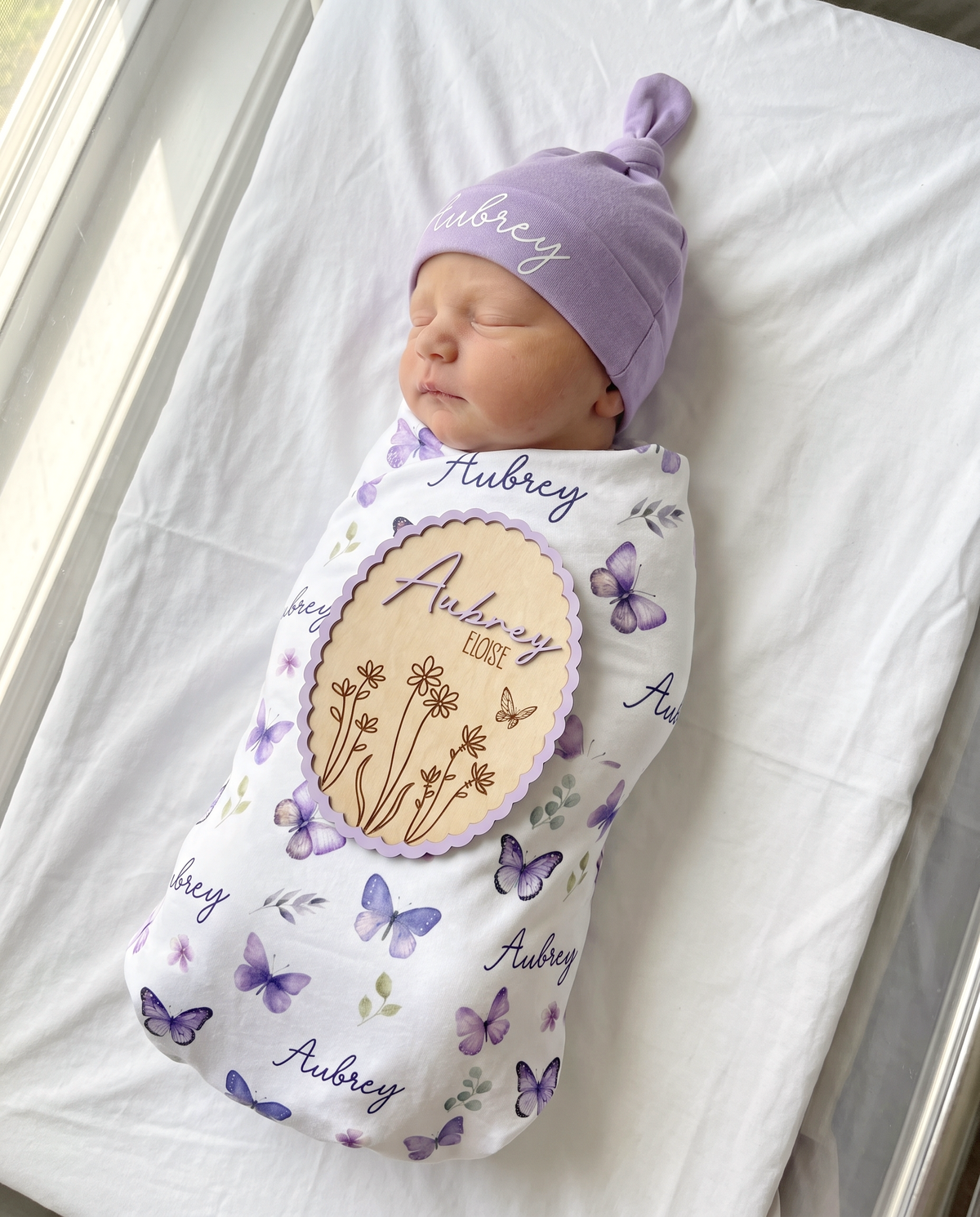 Personalized wood announcement sign in Scalloped Bloom design, shown from above with a matching swaddle and knotted hat