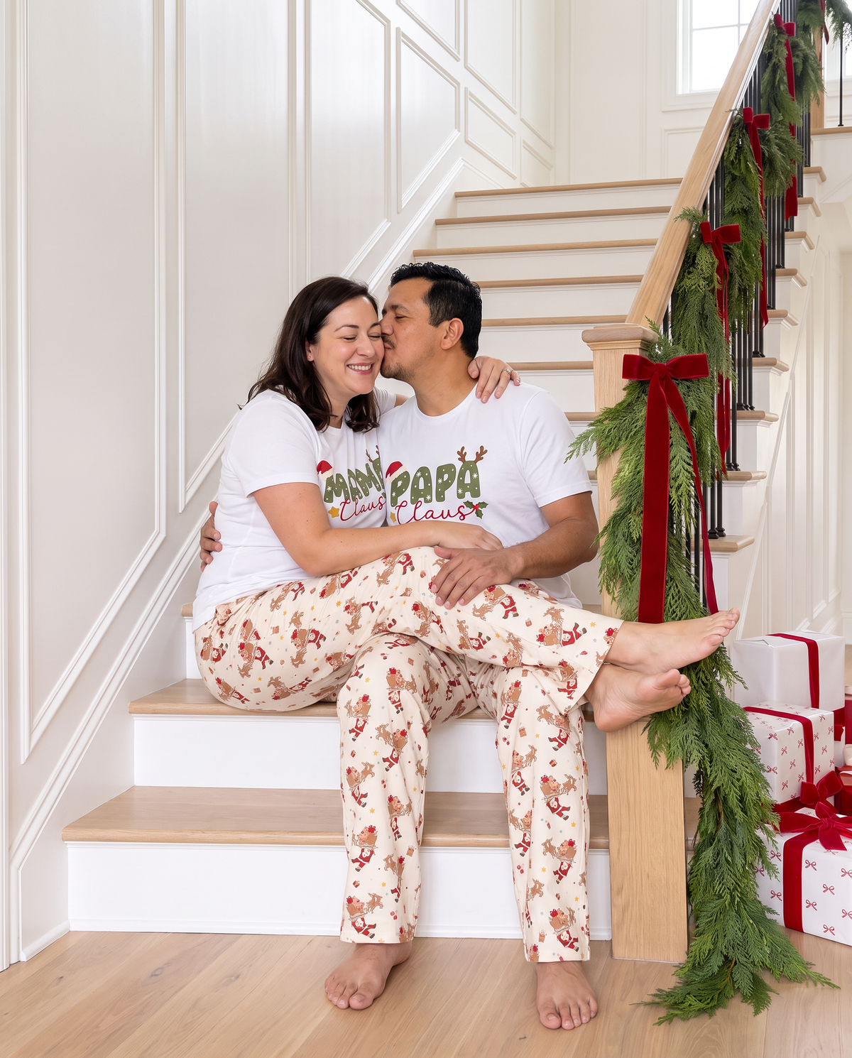 Couple in Santa's Joyride lounge pants sharing a kiss on the garland staircase