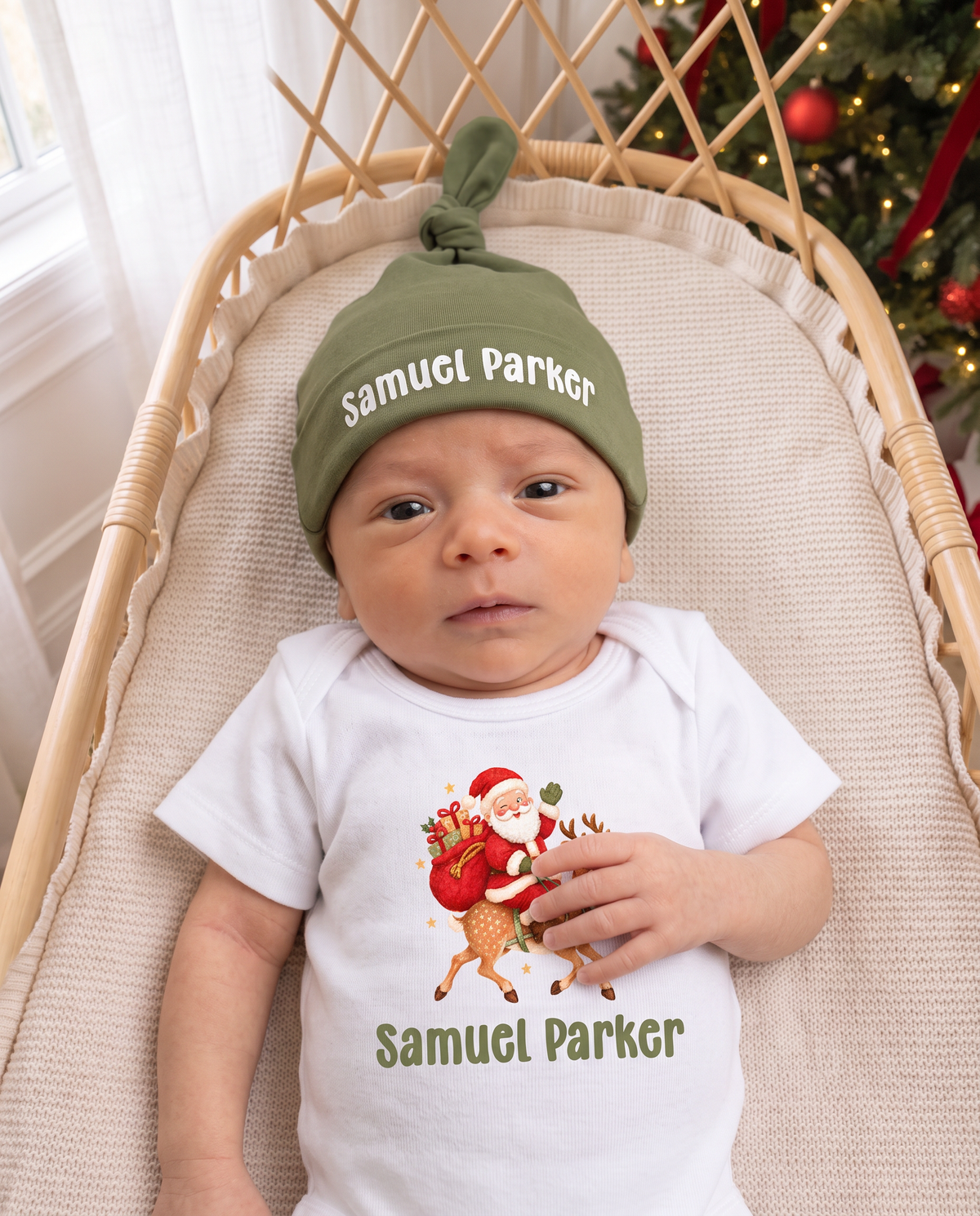Baby in a personalized Santa graphic bodysuit and name knotted hat in a rattan bassinet