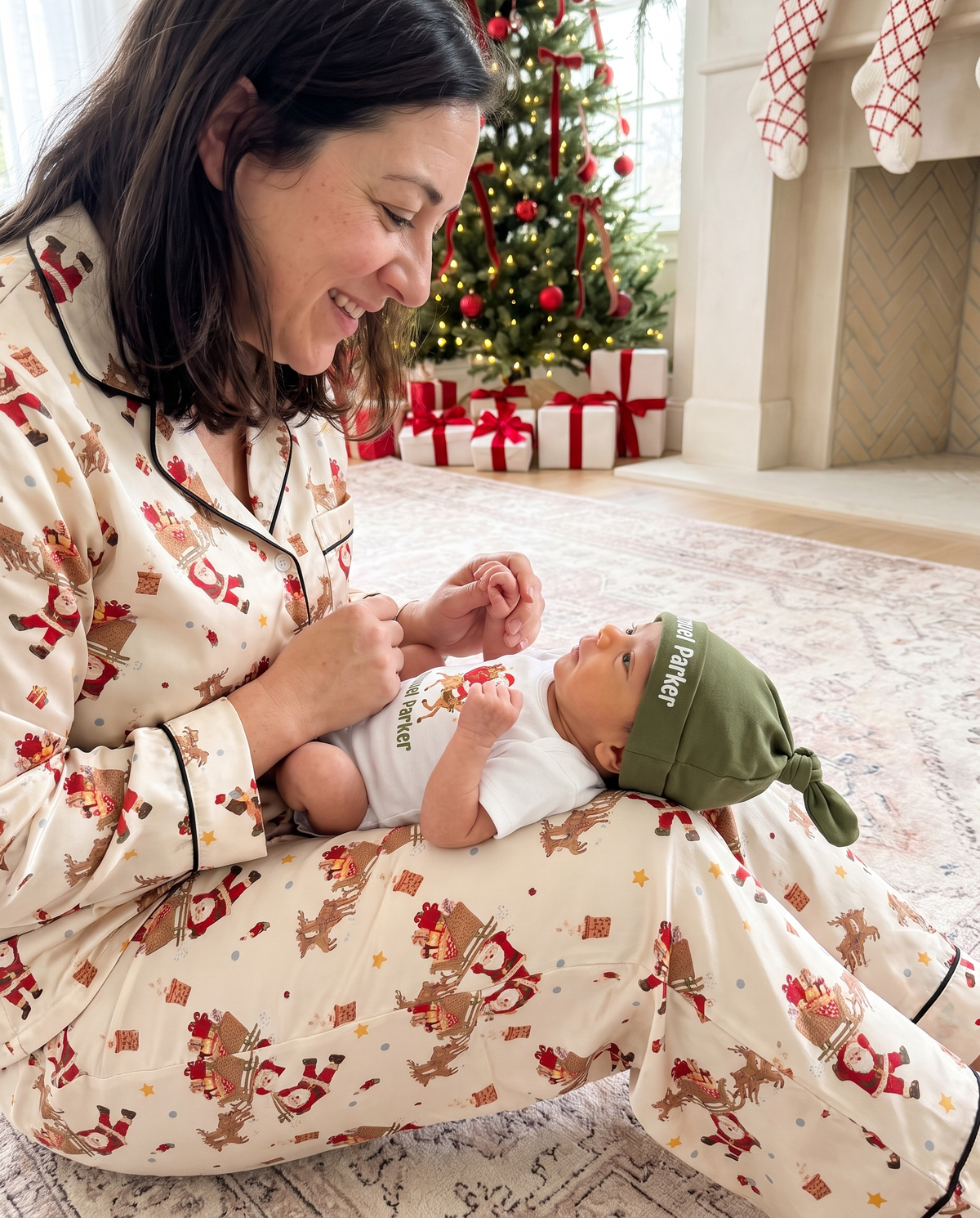 Mom in Santa's Joyride pajamas playing with her baby in the graphic bodysuit on the rug