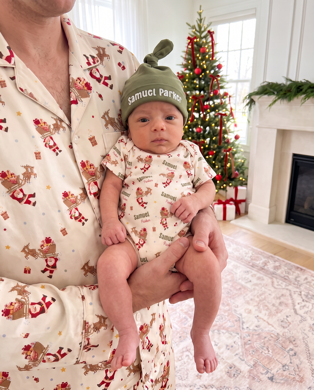 Parent holding a baby in the personalized Santa's Joyride bodysuit and name knotted hat by the Christmas tree