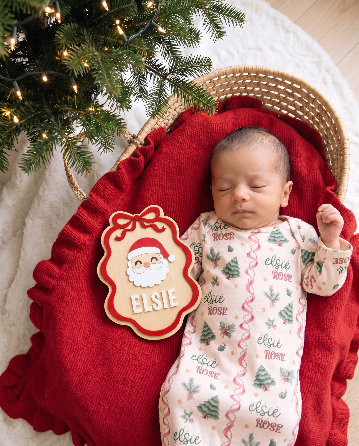 Ribbon Santa personalized wood announcement sign lying on a red ruffle blanket beside a sleeping baby in a woven bassinet
