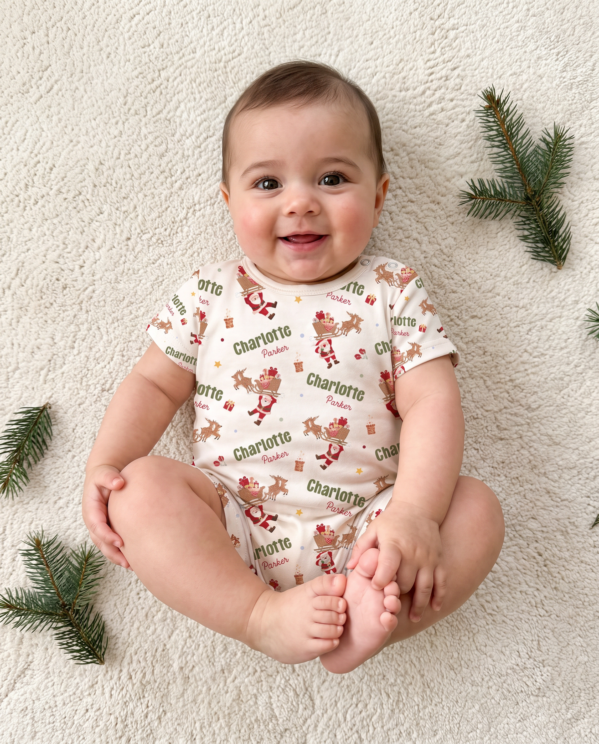 Baby sitting in Santa's Joyride short-sleeve snap romper on a cream blanket with pine sprigs